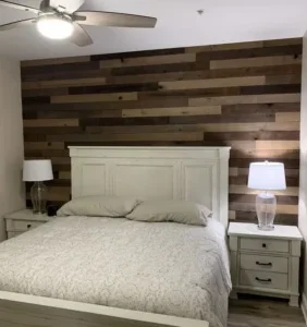 A bedroom with a white bed and matching nightstands, each with a clear glass lamp. The accent wall features horizontal wooden planks in varying browns.