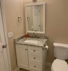 Small bathroom with a white vanity and granite countertop, a sink with a mirror above, a white toilet, beige walls, and a door partially visible to the left.