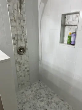 Modern shower with white and light gray tile, featuring a vertical mosaic accent, a mounted handheld showerhead, and a built-in wall niche for bottles.