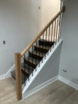 A small staircase with a wooden handrail and black balusters, set against a light gray wall, with tiled flooring at the base and unfinished steps.