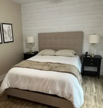 A neatly made bed with a beige upholstered headboard and white bedding sits between two black nightstands with lamps. Behind the bed is a white shiplap accent wall. Three framed botanical prints hang on the tan wall above wood flooring.