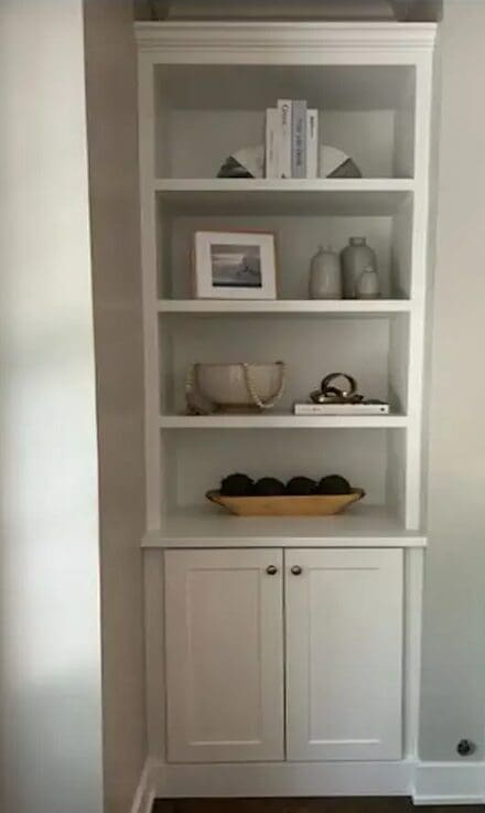 White built-in bookshelf with three shelves displaying books, decor items, vases, a framed photo, decorative spheres in a bowl, and closed cabinets at the bottom. The shelf is against a light-colored wall.